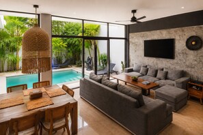 Ein modernes Wohnzimmer mit Blick auf einen Pool, grauem Sofa, einem Holzesstisch und einem Deckenventilator.