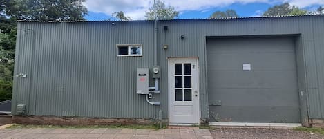 Il s’agit d’un bâtiment industriel d’un seul étage, doté d’une façade en métal ondulé gris, d’une porte blanche avec une fenêtre et d’une grande porte de garage grise.