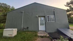Um edifício metálico térreo com porta e janela, rodeado por grama e árvores.
