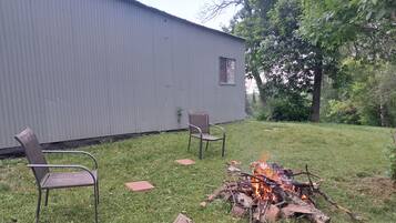 A grassy outdoor area with a fire pit, two chairs, and a building in the background.