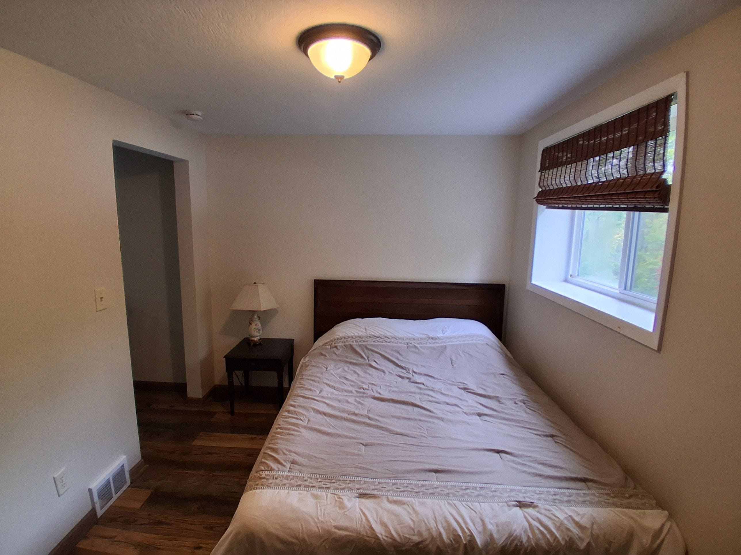 Ein Schlafzimmer mit einem Einzelbett, einem Nachttisch mit Lampe, einem Fenster mit Jalousien und einer Tür zu einem anderen Raum.