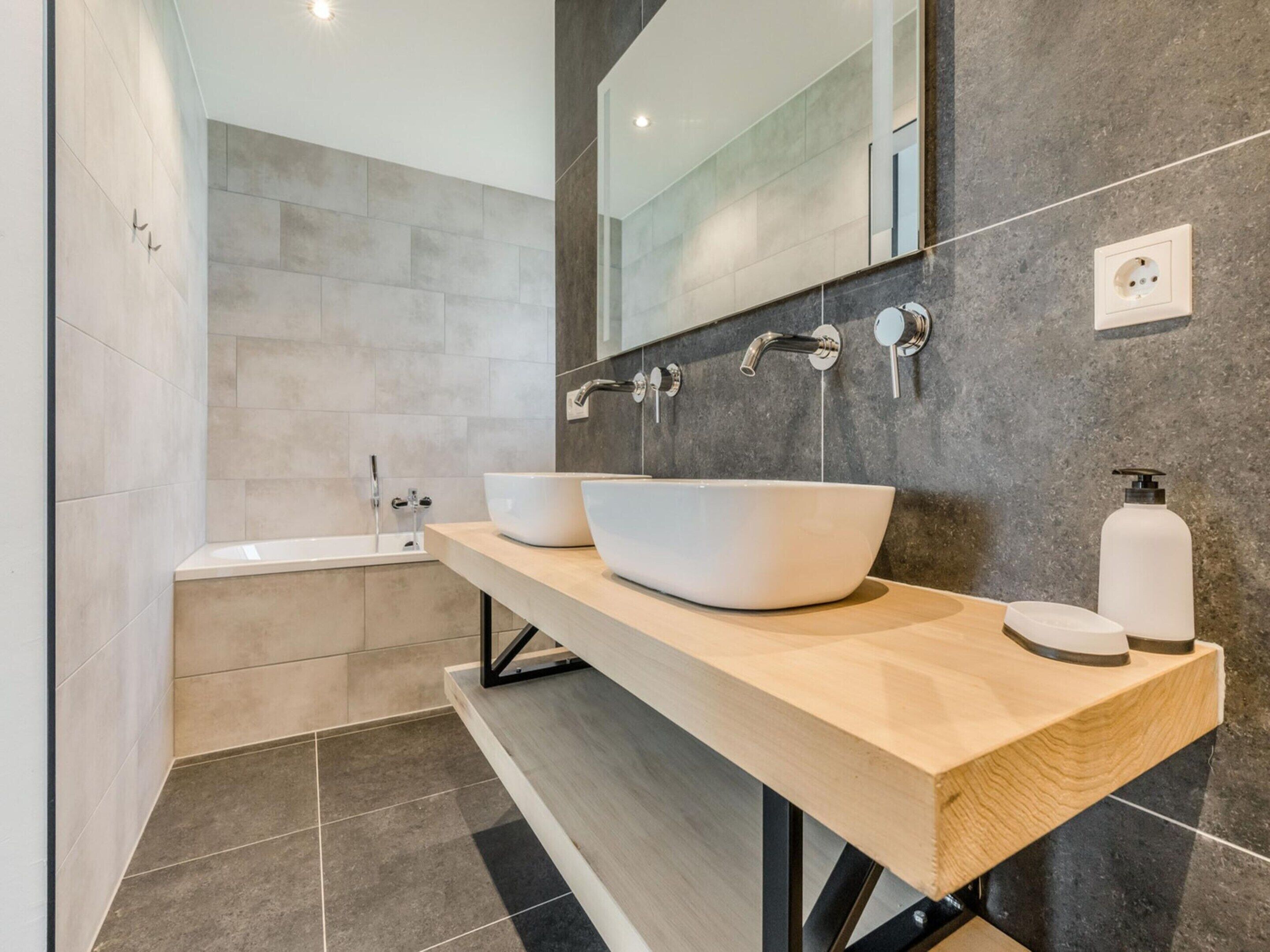 A modern bathroom with a wooden vanity, two white vessel sinks, a large mirror, and a bathtub.