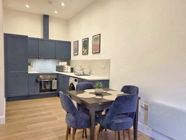 A modern kitchen with grey cabinets, a built-in oven, and a washing machine. There is a dining table with blue chairs and a small potted plant. - Louisville Flat 4 in Newmarket (Newmarket)