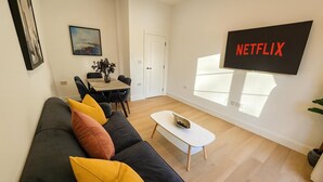 A modern living room with a black sofa, yellow cushions, a white coffee table, and a large TV screen displaying "NETFLIX". - Louisville Flat 3 in Newmarket (Newmarket)