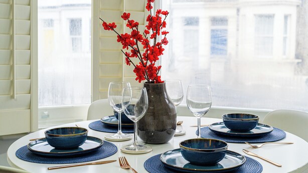 Ein Esstisch ist gedeckt mit blauen Tellern, Schüsseln und Besteck, eine Vase mit roten Blumen und ein Fenster mit Jalousien.