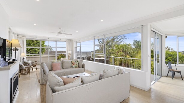 A spacious living area with a sofa, chairs, a TV, and a balcony with large windows. - Spacious 5-bed Beach House With Ocean View (Sydney)