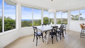 A dining area with a large table and chairs, large windows offering a view of greenery and the sea. - Spacious 5-bed Beach House With Ocean View (Sydney)