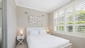 A bedroom with a bed, two bedside tables with lamps, a wall-mounted artwork, and a window with blinds. - Spacious 5-bed Beach House With Ocean View (Sydney)