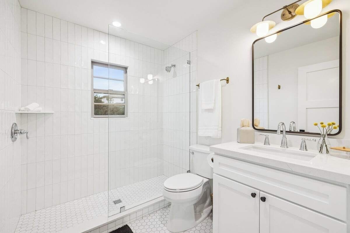 A modern bathroom with white tiles, a glass shower enclosure, a toilet, a vanity with a mirror, and a window with a view.