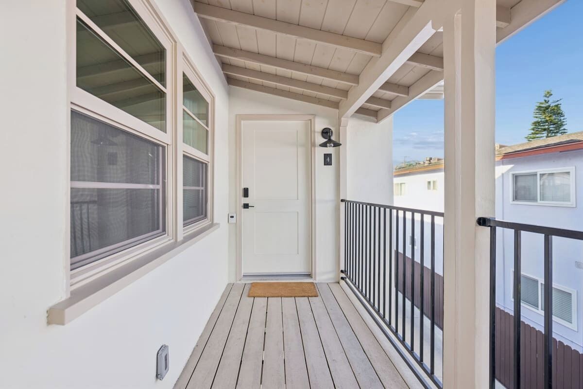 A covered balcony with a wooden floor, a white door, and a black railing.