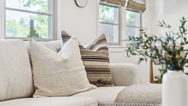 Ein beiges Sofa mit mehreren Kissen, eine weiße Vase mit Grünpflanzen und ein Fenster mit Jalousien.