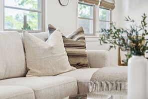 A beige sofa with multiple cushions, a white vase with greenery, and a window with blinds. - Casa Yano by The Beach (Santa Barbara)