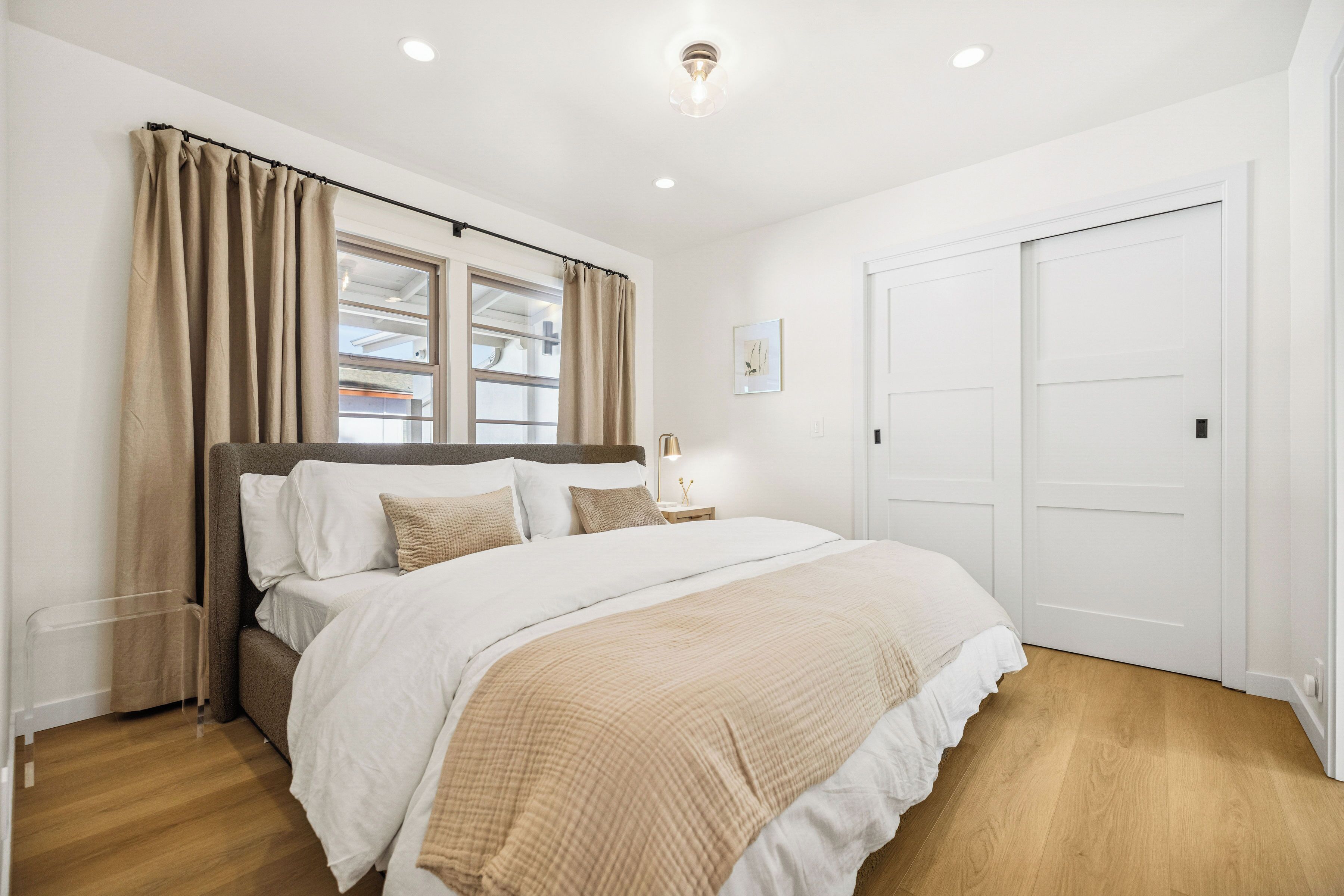 A bedroom with a large bed, beige curtains, a bedside table with a lamp, and a white door.