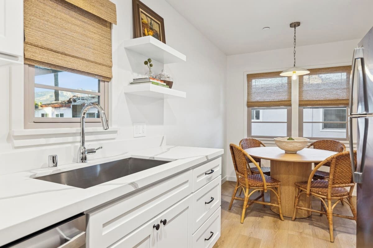 A modern kitchen with a white countertop, stainless steel sink, and a dining area with wicker chairs and a round table.