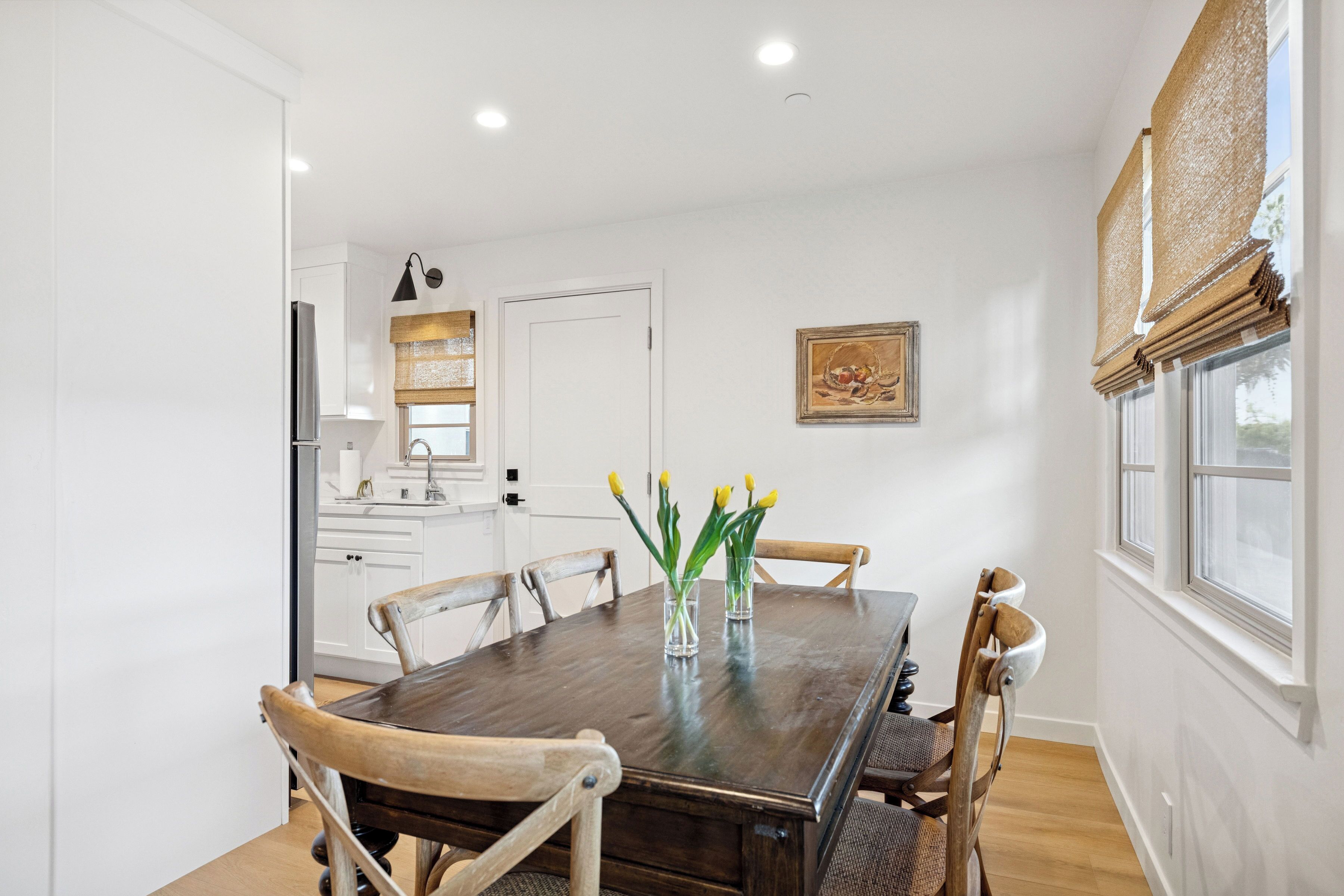 A dining area with a wooden table, chairs, and a vase with flowers. There is a kitchen in the background with white cabinets and a refrigerator.