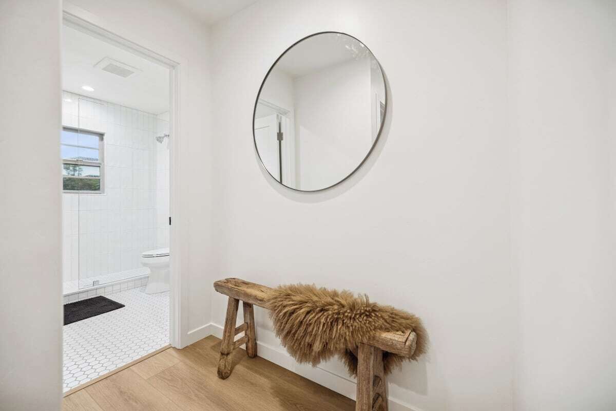Een badkamer met een vloer van witte tegels, een houten bankje met een bont plaid en een ronde spiegel aan de muur.