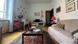 A living room with a sofa, a coffee table, a TV, a red chair, a patterned rug, and a door leading to another room. - Central and spacious apartment 100m from the sea! (Alassio)