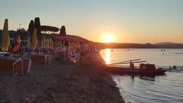 Une plage avec des parasols et des tables, un bateau nommé « SALVATAGGIO » sur l’eau, et un coucher de soleil en arrière-plan.