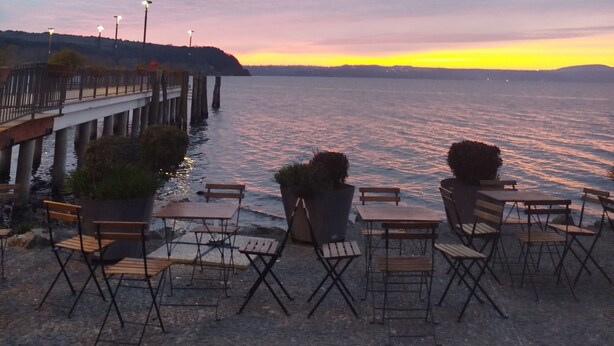 A waterfront seating area with tables and chairs, a pier with street lamps, and a sunset view. - Gabry House - sunset on the lake (anguillara sabazia)