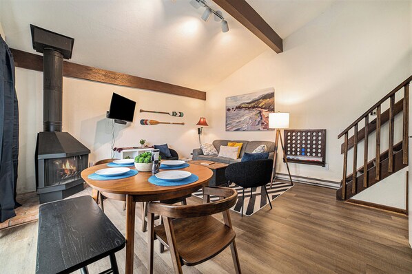 Una sala de estar acogedora con chimenea, una mesa de comedor preparada para cuatro comensales y una escalera que sube a un nivel superior.