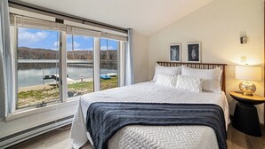 A bedroom with a large window overlooking a lake, a bed with white bedding, and a nightstand with a lamp. - Serene Northern Michigan 3-bedroom townhome near Boyne Mountain and Gaylord (Gaylord)