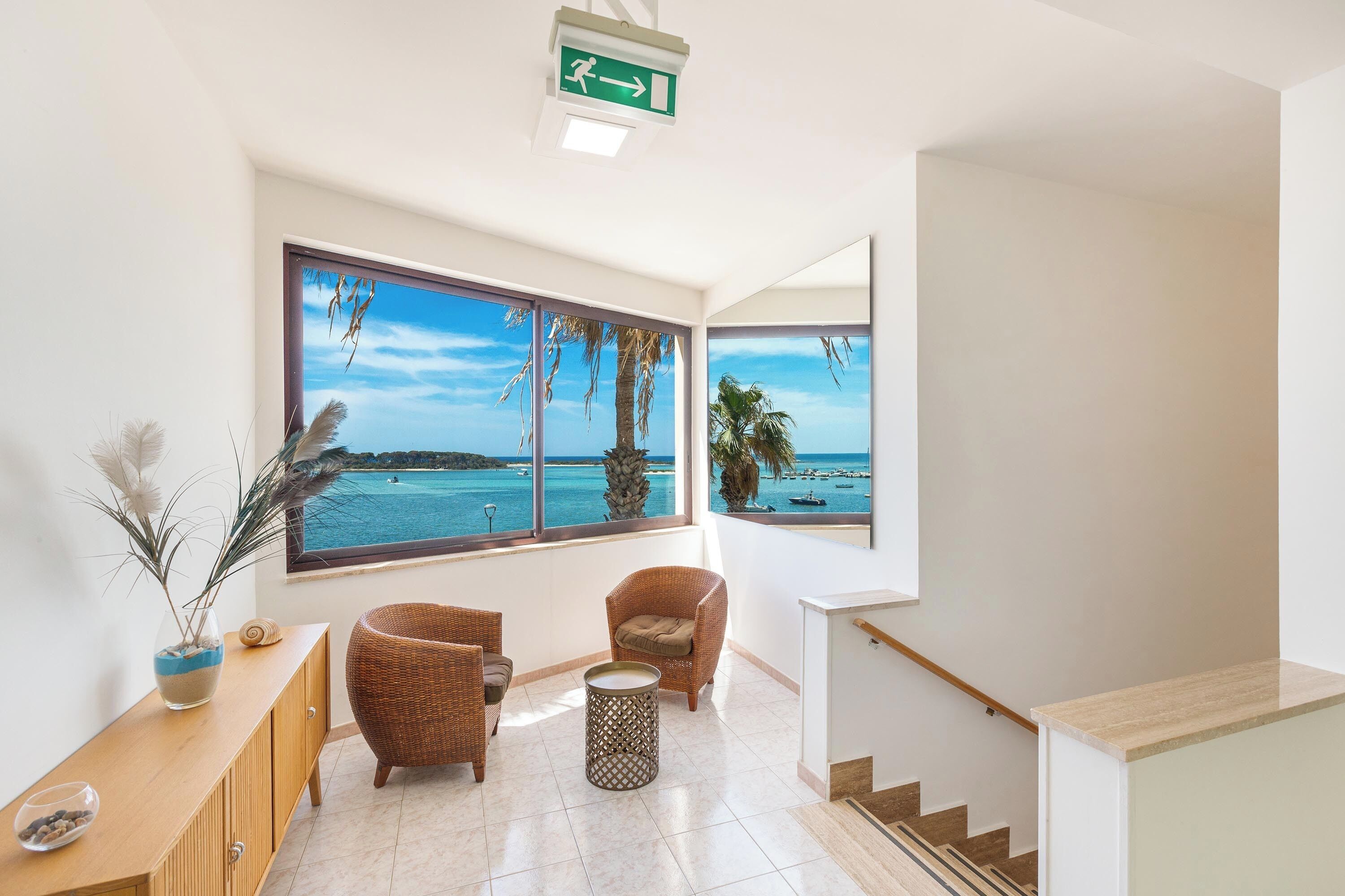 Ein geräumiges Zimmer mit großem Fenster und Blick auf das Meer und Boote, ein Holzkonsolentisch, zwei Korbstühle und ein kleiner runder Tisch.