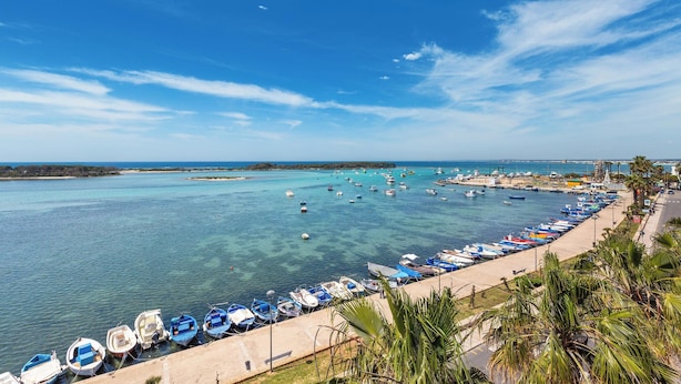 A coastal marina with numerous boats docked along a paved walkway, overlooking a clear blue sea.