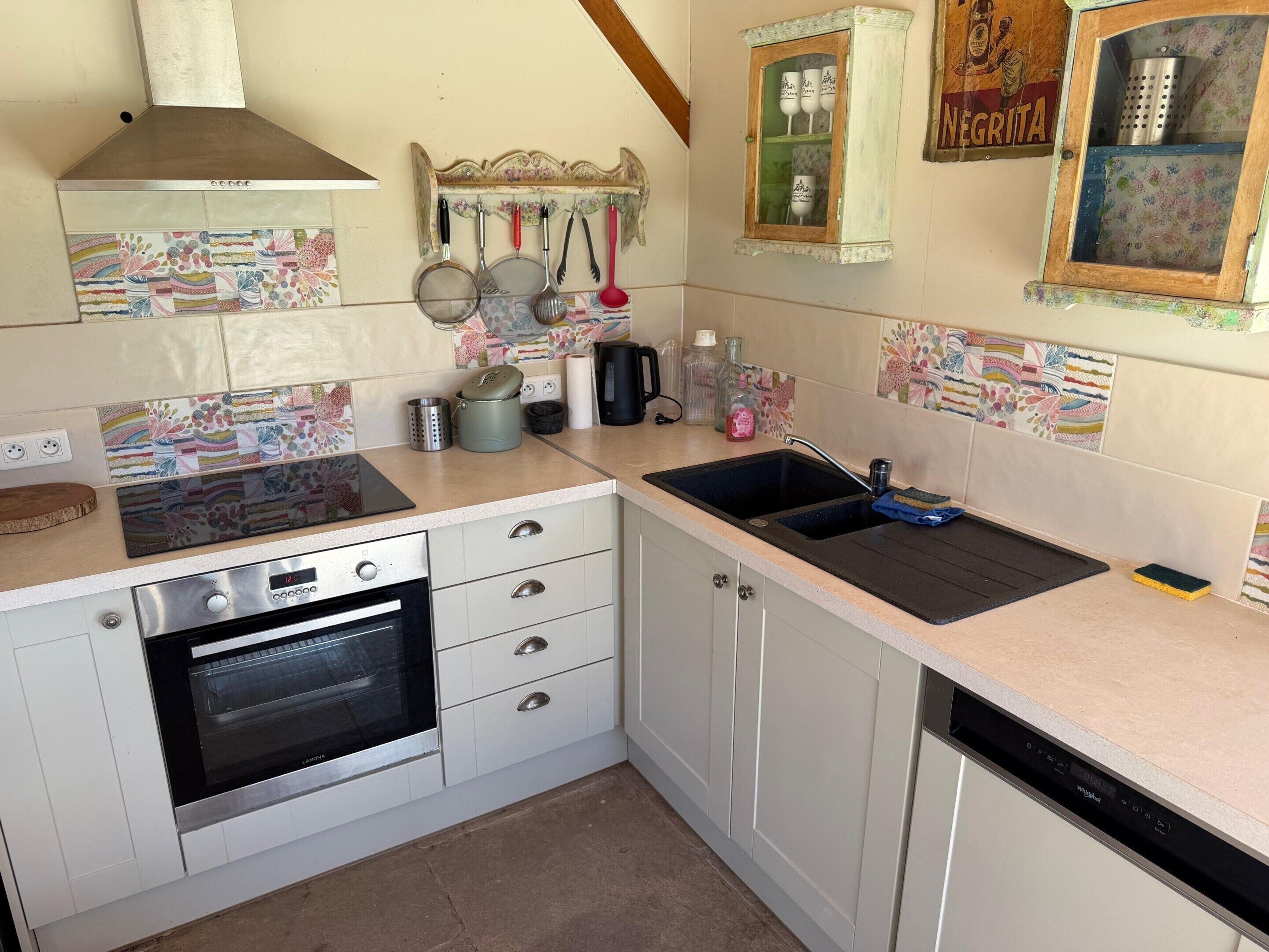 A compact kitchen with a built-in oven, a sink, and a dishwasher. There are decorative tiles on the backsplash and a small cabinet with glassware.