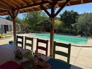 Ein überdachter Außenbereich mit Pool, Holzstühlen, einem Tisch mit Flasche und Gläsern sowie Blick auf Bäume und ein Gebäude.