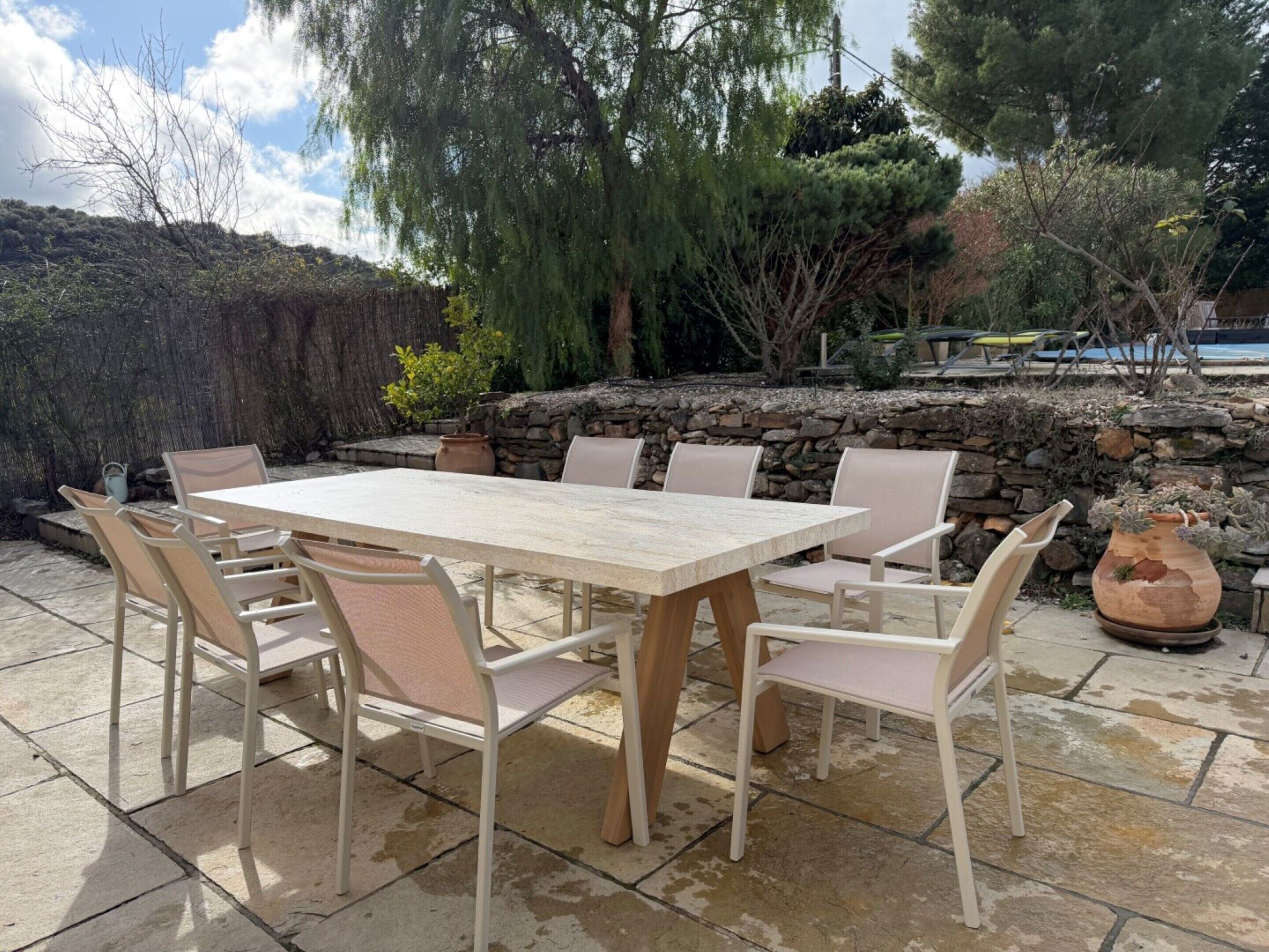 Un patio al aire libre con una mesa y sillas de piedra, rodeado por un muro de piedra y diversas plantas.
