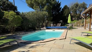 Μια ορθογώνια πισίνα με πλακόστρωτο χώρο γύρω της, μια σκεπαστή περιοχή με καθίσματα και καρέκλες, και μια πέργκολα με μια πράσινη ομπρέλα.