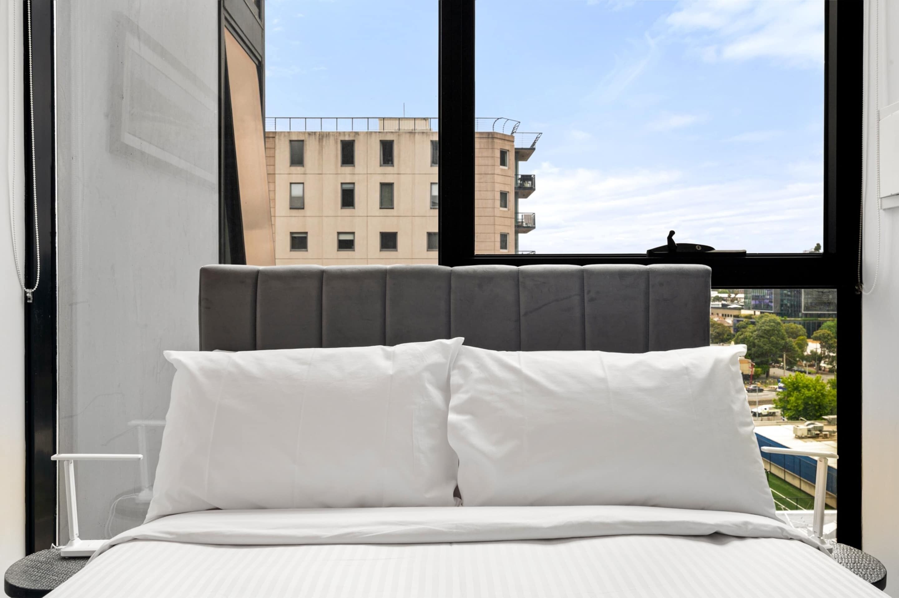 Ein Bett mit weißer Bettwäsche und einem grauen Kopfteil, ein Fenster mit Blick auf Gebäude und einen klaren Himmel.