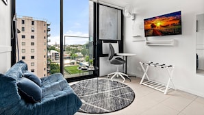 A modern living room with a large window, a sofa, a small round rug, a side table, a chair, a TV, and a drying rack. - South Melbourne Apartment With Gym Access (Melbourne)