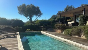 Ein klares Schwimmbecken mit Holzterrasse und Liegestühlen, umgeben von Grünflächen und einem Haus.