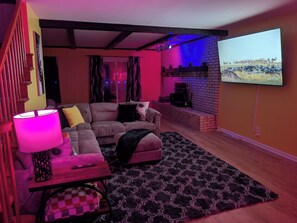 A living room with a sofa, a table with a lamp, a TV mounted on the wall, and a brick fireplace. - 5BR Home, Pool + Game Room | Sleeps 16+ (Evansville)