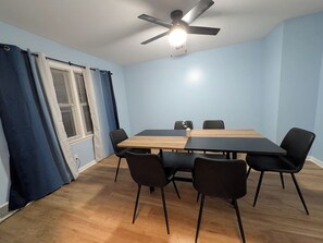 A room with a ceiling fan, a table with chairs, a window with blinds, and a curtain.