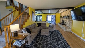 A living room with a sectional sofa, a wooden staircase, a fireplace, and a TV mounted on the wall. - 5BR Home, Pool + Game Room -- Sleeps 16+ (Evansville)