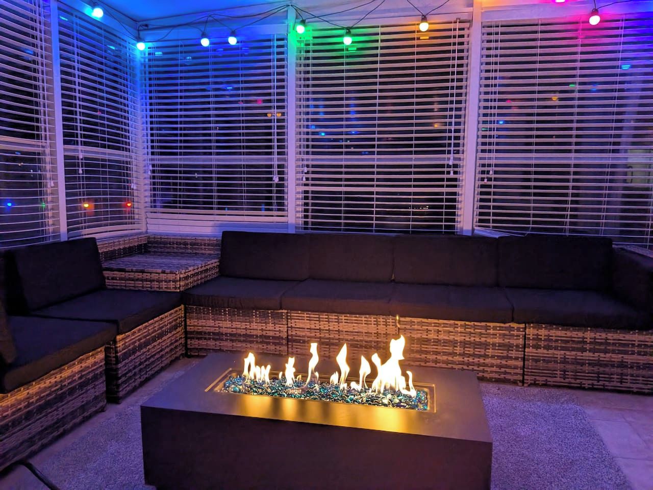 An outdoor seating area with a fire feature, cushioned seating, and a view through window blinds.
