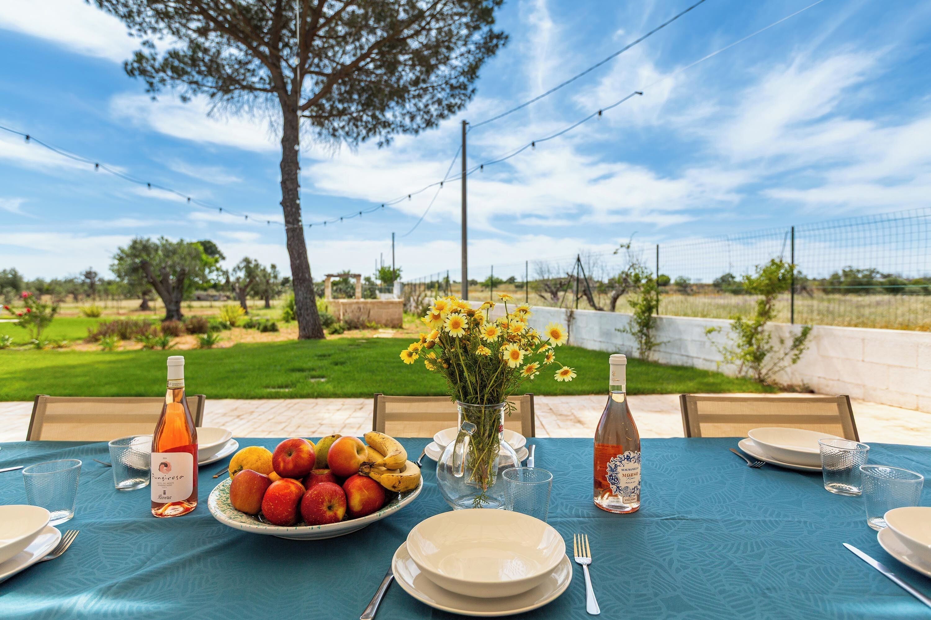 An outdoor dining table set with plates, cutlery, a bottle of rosé wine, a bottle of white wine, a vase with yellow flowers, and a plate of fruit.