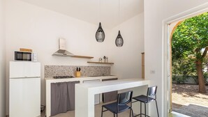 Cocina compacta con refrigerador blanco, microondas y estufa, una pequeña barra con dos sillas, y vista a un jardín desde un vano abierto.