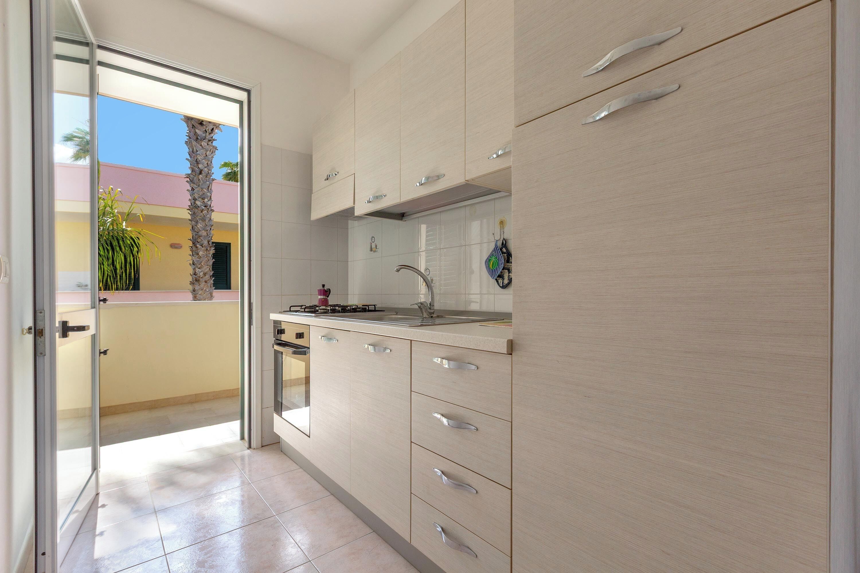 Une cuisine moderne avec des meubles de couleur bois clair, un évier en acier inoxydable et une vue sur un balcon agrémenté de palmiers.