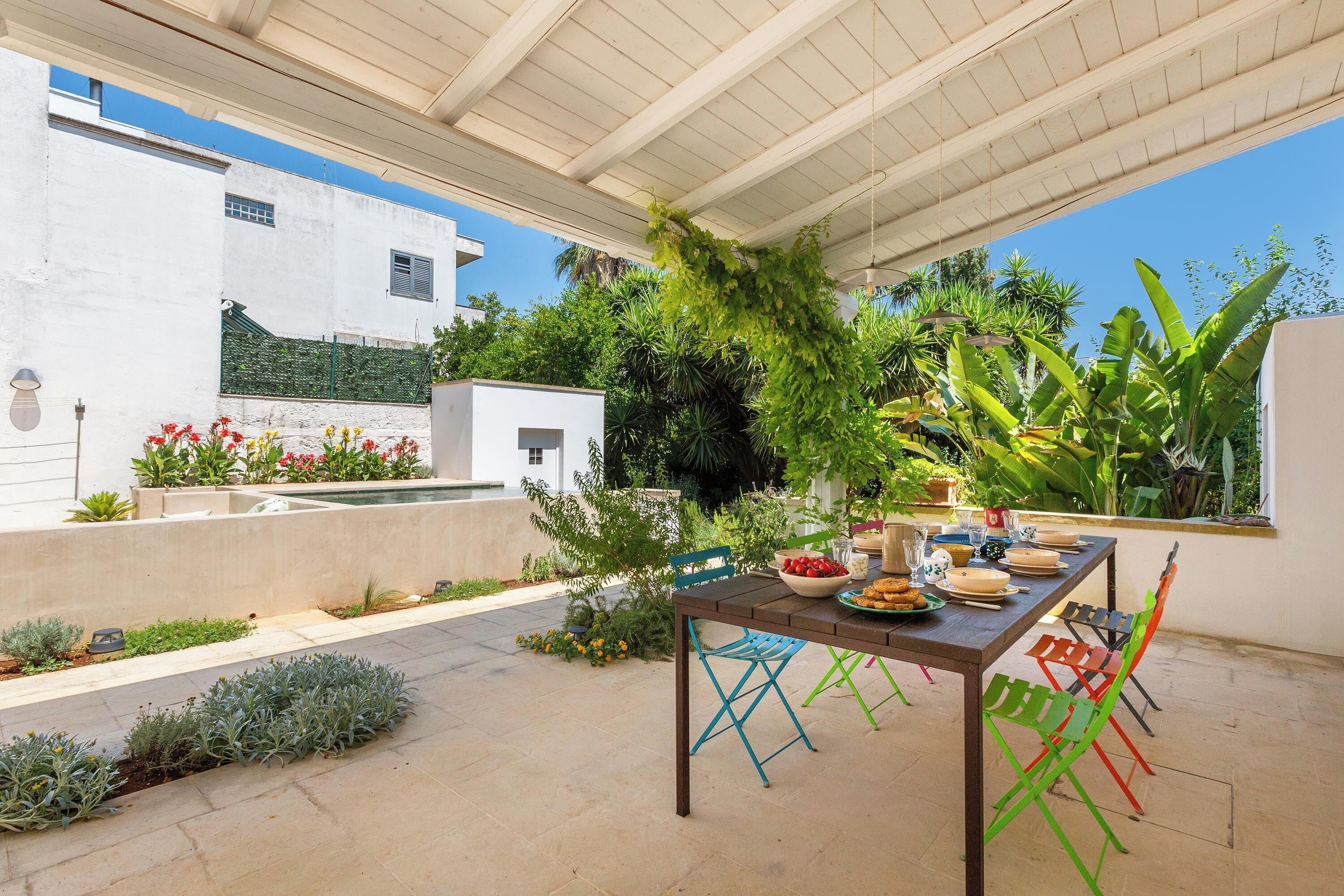 Un área de comedor al aire libre con una mesa lista para dos personas, rodeada de sillas de colores, bajo una pérgola con plantas trepadoras.