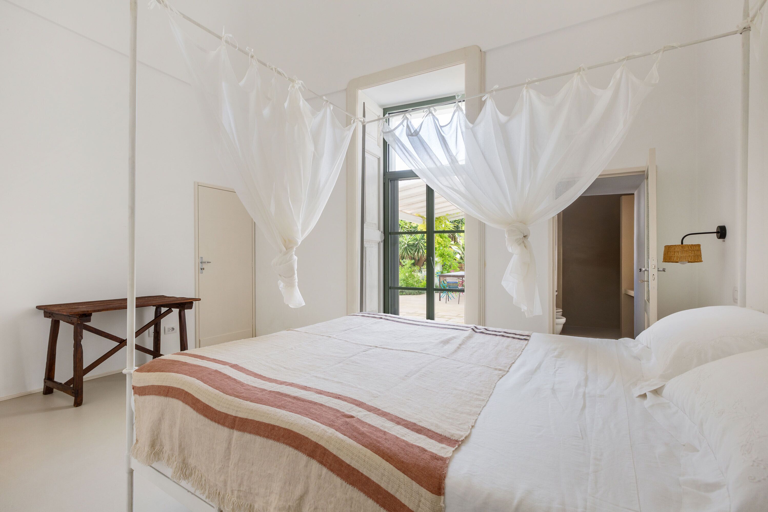 A bedroom with a bed, a wooden table, and a window with a view of greenery.