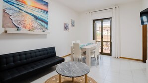 Un saló modern amb un sofà negre, una taula de centre de marbre i una zona de menjador amb cadires blanques i una taula. Un quadre gran d'un capvespre sobre l'oceà adorna la paret.