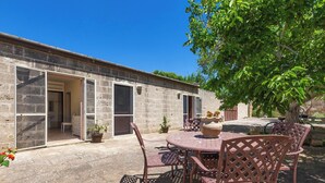Une terrasse extérieure avec une table ronde et des chaises, un bâtiment en pierre dont les portes sont ouvertes, et un arbre qui offre de l’ombre.