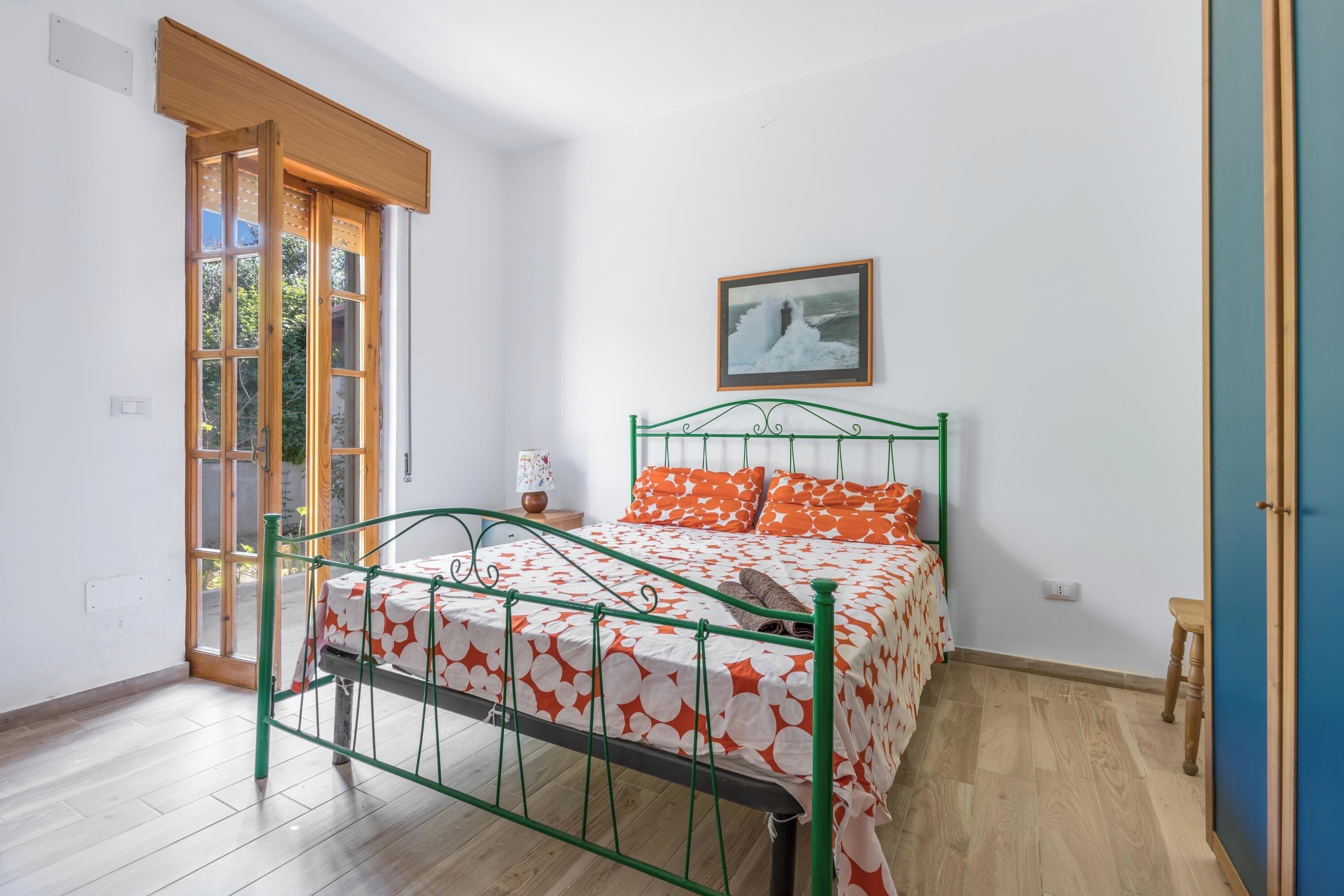 Une chambre avec un lit en métal vert, une couvre-lit à motifs blancs et orange, un tableau encadré au mur et une porte en bois avec un panneau de verre.