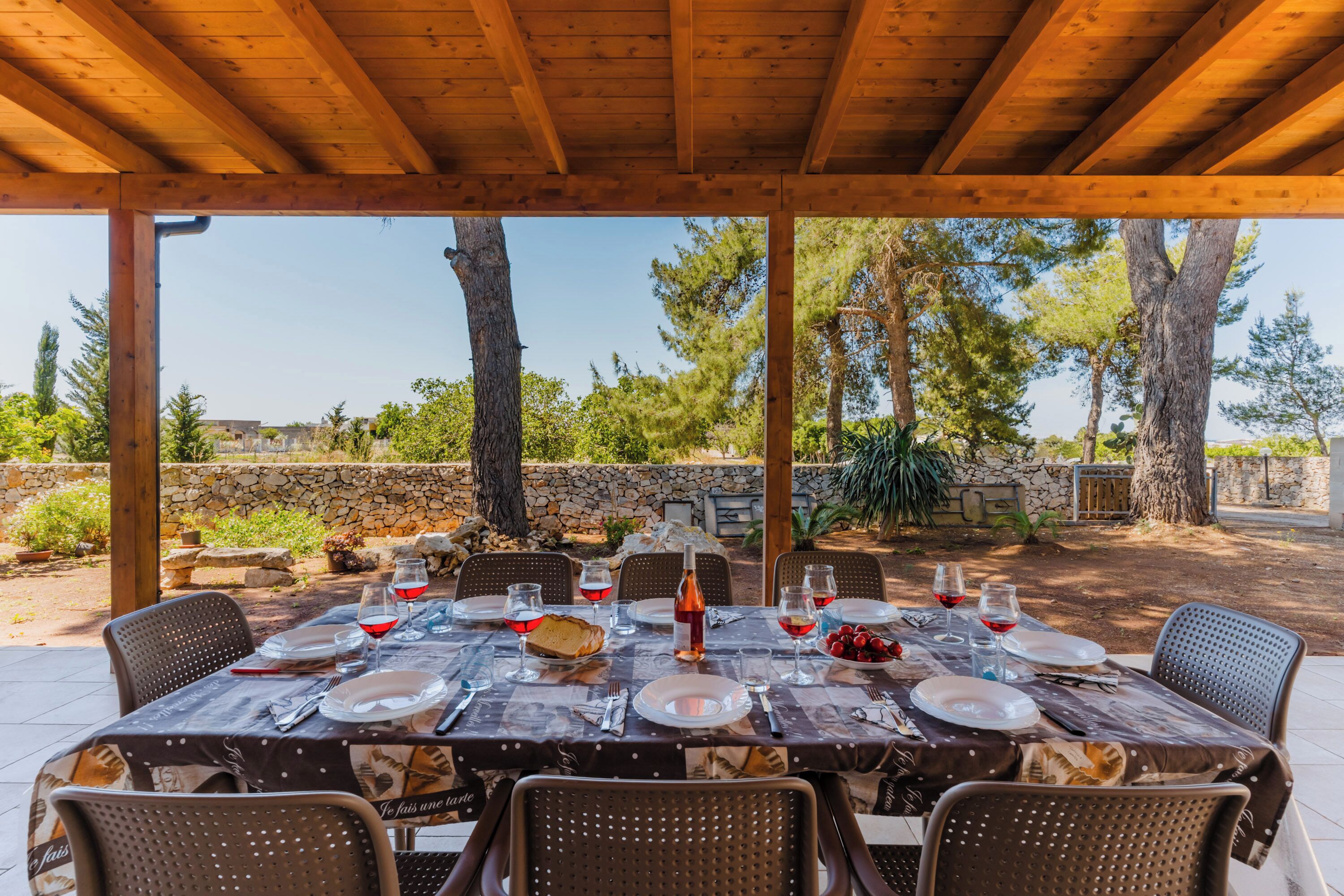 An outdoor dining area with a wooden pergola, stone wall, and trees in the background.