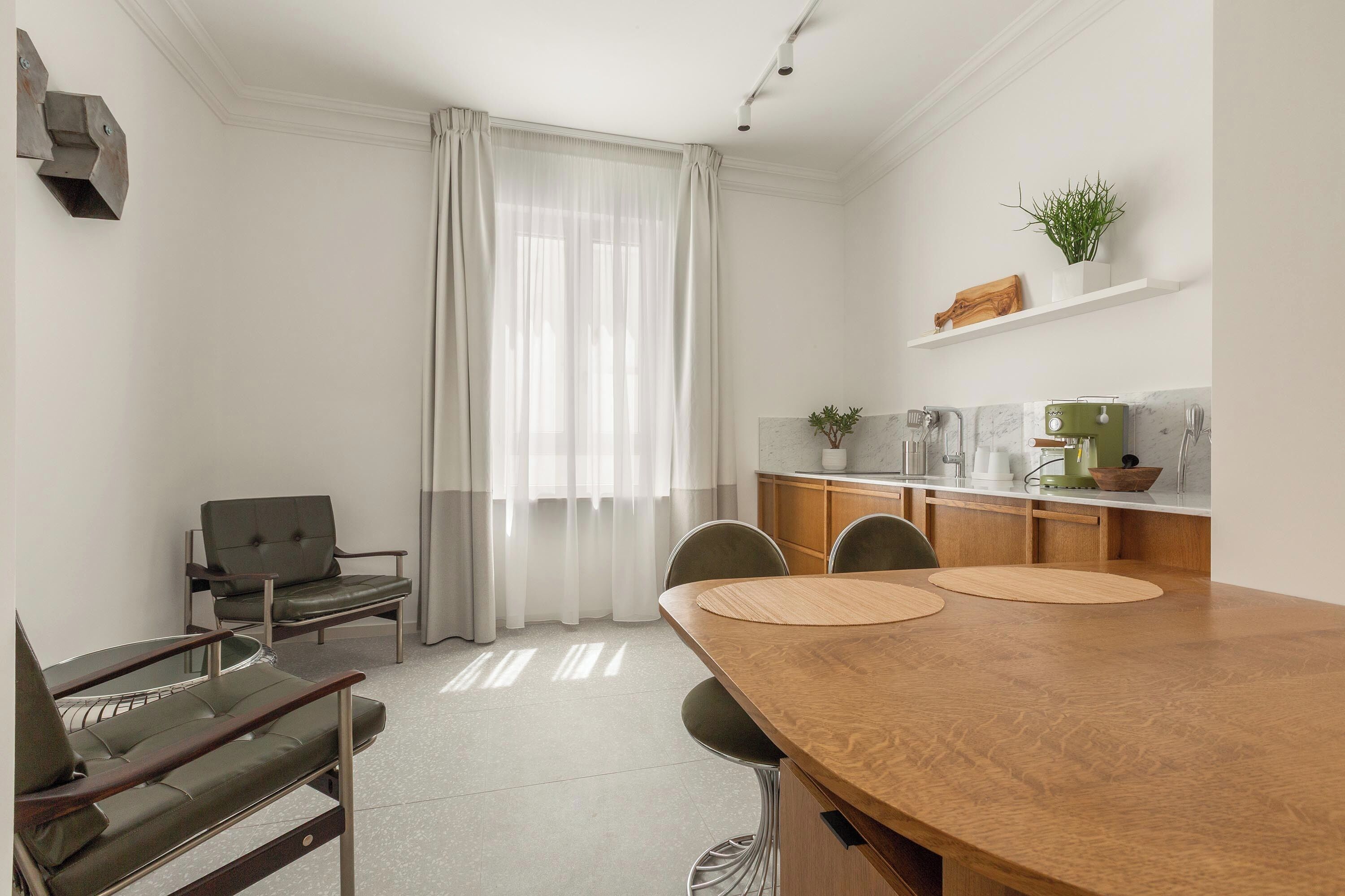 A modern, well-lit room with a wooden dining table, two chairs, a sofa, and a small kitchen area with a coffee machine.