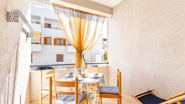 Un balcon avec une table dressée pour deux personnes, des chaises en bois avec des coussins, un petit hamac et une vue sur un immeuble doté de balcons.