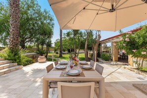 Un área de comedor al aire libre con una mesa dispuesta para cuatro personas, rodeada de sillas y cubierta por una sombrilla grande.